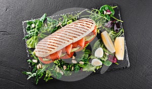 Sandwich with red fish, eggs, avocado, fresh vegetables and greens on black shale board over black stone background