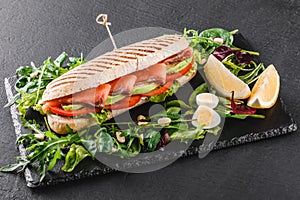 Sandwich with red fish, eggs, avocado, fresh vegetables and greens on black shale board over black stone background