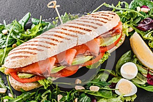 Sandwich with red fish, eggs, avocado, fresh vegetables and greens on black shale board over black stone background