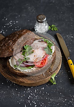 Sandwich with roasted pork, soft cheese, tomatoes and arugula on the wooden cutting board on dark background.