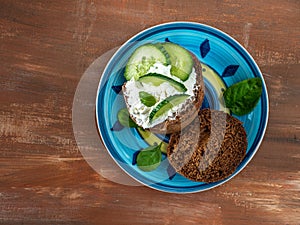 Sandwich on a rye bun with soft cheese and cucumber with basil leaves on a colored plate