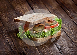 Sandwich with smoked meat, green salad, fresh tomatoes and pickled cucumbers, on kitchen wooden table background