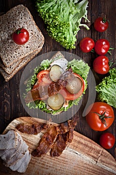 Sandwich with smoked meat, green salad, fresh tomatoes and pickled cucumbers, on kitchen wooden table background