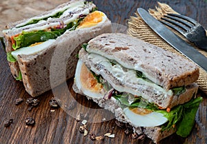 Sandwich stuffed with chicken breast, cheese, boiled eggs and fresh vegetables serve with fork and knife on wooden plate