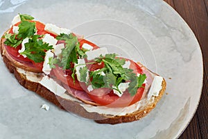 Sandwich of toast and rye bread with greens, tomatoes, feta cheese and ham in grey plate on dark wooden background.