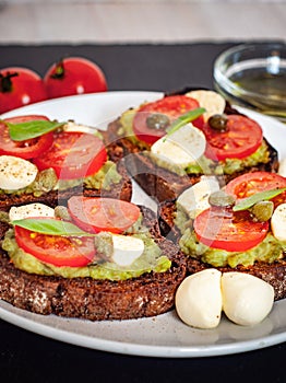 Sandwich toasts with tomatoes cherry, mozzarella, avocado, basil and olive oil. Side view on a dark stone dish.