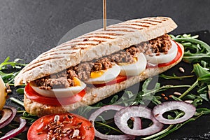 Sandwich with tuna, eggs, fresh vegetables and greens on black shale board over black stone background.