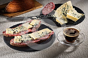 Sandwiches with cheese of Dorblu and raw smoked sausage on black slate plates, a cheese knife and a cup of hot coffee