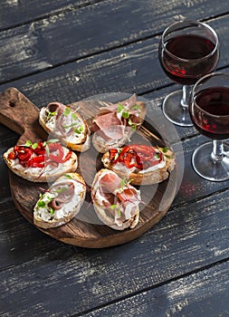 Sandwiches with goat cheese, anchovies, roasted peppers, ham and two glasses of red wine on a wooden rustic board.