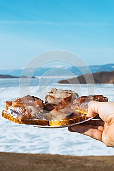 Sandwiches with omul on a white round plate. A plate of sandwiches in hands. Winter Baikal in ice. Fish dishes.