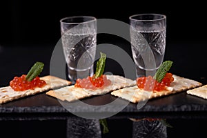 Sandwiches with red caviar with butter and a leaf of mint from cracker with glasses of vodka on a slate plate on a dark background
