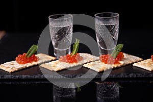 Sandwiches with red caviar with butter and a leaf of mint from cracker with glasses of vodka on a slate plate on a dark background