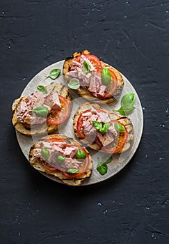 Sandwiches with tomatoes, basil and tuna fish on wooden cutting board on dark background, top view. Healthy breakfast, tapas