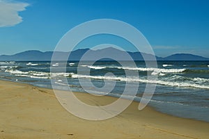 Sandy beach, blue sea with white sea foam and mountains on blue sky background