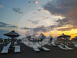 Sandy beach white lounge chairs aligned next to sea for relaxation with parasols and sunshades during sunrise with colorful sky