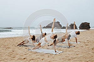 Sandy mat sessions. Group of young calm men and women enjoying morning yoga, doing lateral side plank on the beach