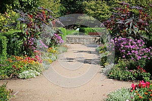 a sandy path in a flower park and at the end of the path there is a brick fountain