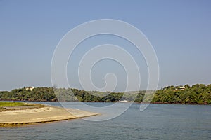 A sandy shore on the background of the river and hills with green trees and houses under a clear blue sky