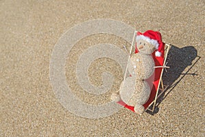 Sandy snowman in santa hat sunbathing in beach lounge.