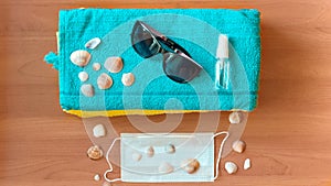 Sanitizer bottle, face mask, sunglasses and shells on a blue towel against a wood background. The concept of a summer