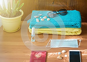 A sanitizer bottle, face mask, sunglasses and shells on a blue towel against a wood background. The concept of a summer vacation