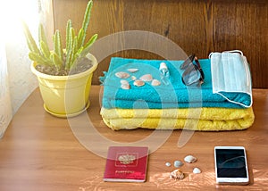 A sanitizer bottle, face mask, sunglasses and shells on a blue towel against a wood background. The concept of a summer vacation