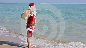 Santa Claus with a bag of gifts on his shoulder walks along the sandy beach