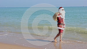 Santa Claus with a bag of gifts on his shoulder walks along the sandy beach