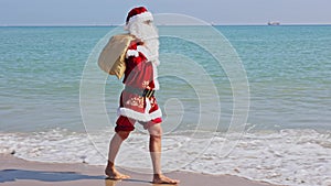 Santa Claus with a bag of gifts on his shoulder walks along the sandy beach