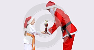 Santa Claus with a black belt is giving little girl in karategi Cup of Karate