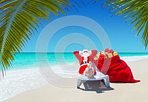 Santa Claus with Christmas sack full of gifts relax on sunlounger at tropical palm beach.