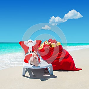 Santa Claus with Christmas sack full of gifts relax on sunlounger barefooted at perfect sandy ocean beach.