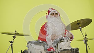 Santa Claus in festive costume with beard and sunglasses plays drums on yellow studio background. Man in a carnival