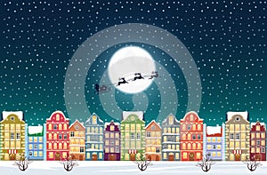Santa Claus flies over a decorated snowy old city town near moon at Christmas eve.