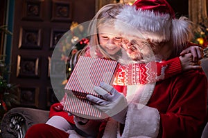 Santa Claus and girl opening Christmas gift near Christmas tree. Child hugging Santa. Magic fulfillment of desires