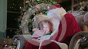 Santa Claus gives magic gift box boy, bright light shines from box to little blonde kid sitting in chair in room with