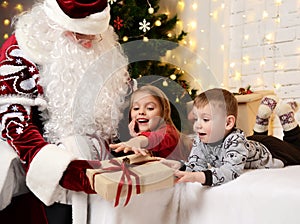 Santa Claus giving present to a happy little cute children boy and girl near Christmas tree