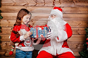 Santa Claus giving a present to a little cute girl near Christmas tree indoor