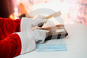 Santa Claus hands holding landline phone and a medical mask over white table background.