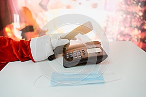 Santa Claus hands holding landline phone and a medical mask over white table background. Online greetings, ordering services for