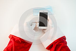 Santa Claus hands holding smart phone and a medical mask over white background. Online greetings, ordering services for Christmas