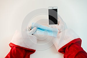 Santa Claus hands holding smart phone and a medical mask over white background. Online greetings, ordering services for Christmas