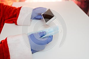 Santa Claus hands holding smart phone and a medical mask over white table background.