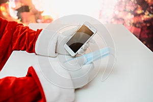 Santa Claus hands holding smart phone and a medical mask over white table background. Online greetings, ordering services for