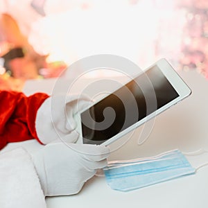 Santa Claus hands holding tablet and a medical mask over white table background. Online greetings, ordering services for Christmas