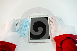 Santa Claus hands holding tablet and a medical mask over white table background. Online greetings, ordering services for Christmas