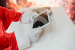 Santa Claus hands holding tablet and a medical mask over white table background. Online greetings, ordering services for Christmas