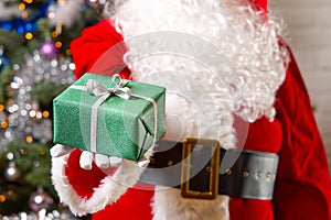 Santa Claus hands holds green gift box opposite christmas tree.