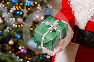 Santa Claus hands holds green gift box opposite christmas tree.