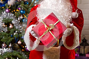 Santa Claus hands holds red gift box opposite christmas tree.